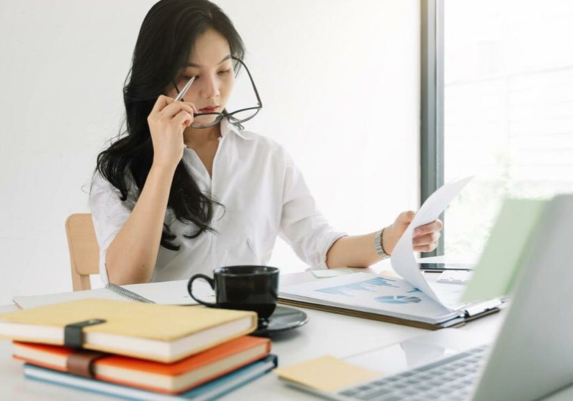 Young asian entrepreneur cogitating thinking making important decision at workplace. Concentrated serious office worker millennial woman analysing results woman getting off her glasses